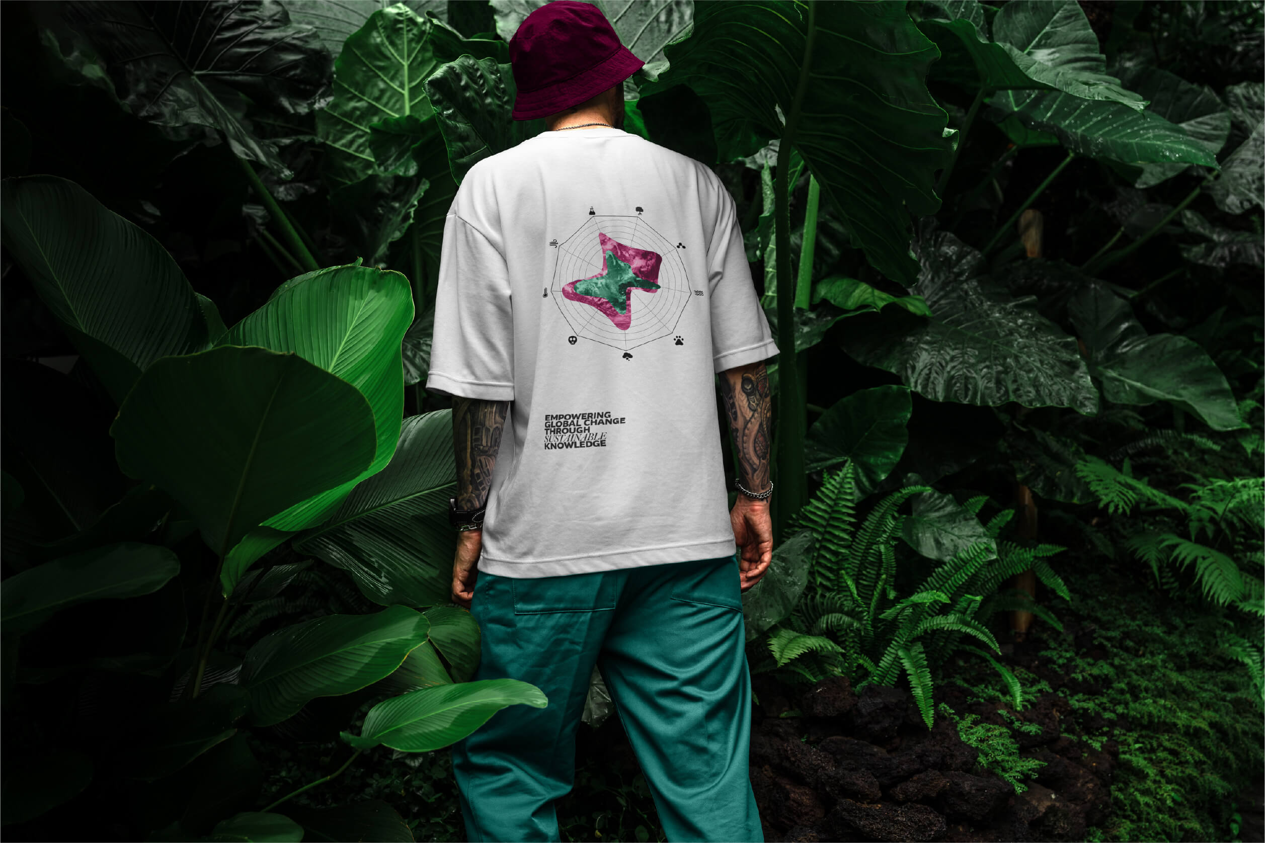 Young man wearing a white t-shirt with a colorful abstract graphic and text reading 'EMPOWERING GLOBAL CHANGE THROUGH SUSTAINABLE KNOWLEDGE' on the back, standing in front of large green tropical plants.