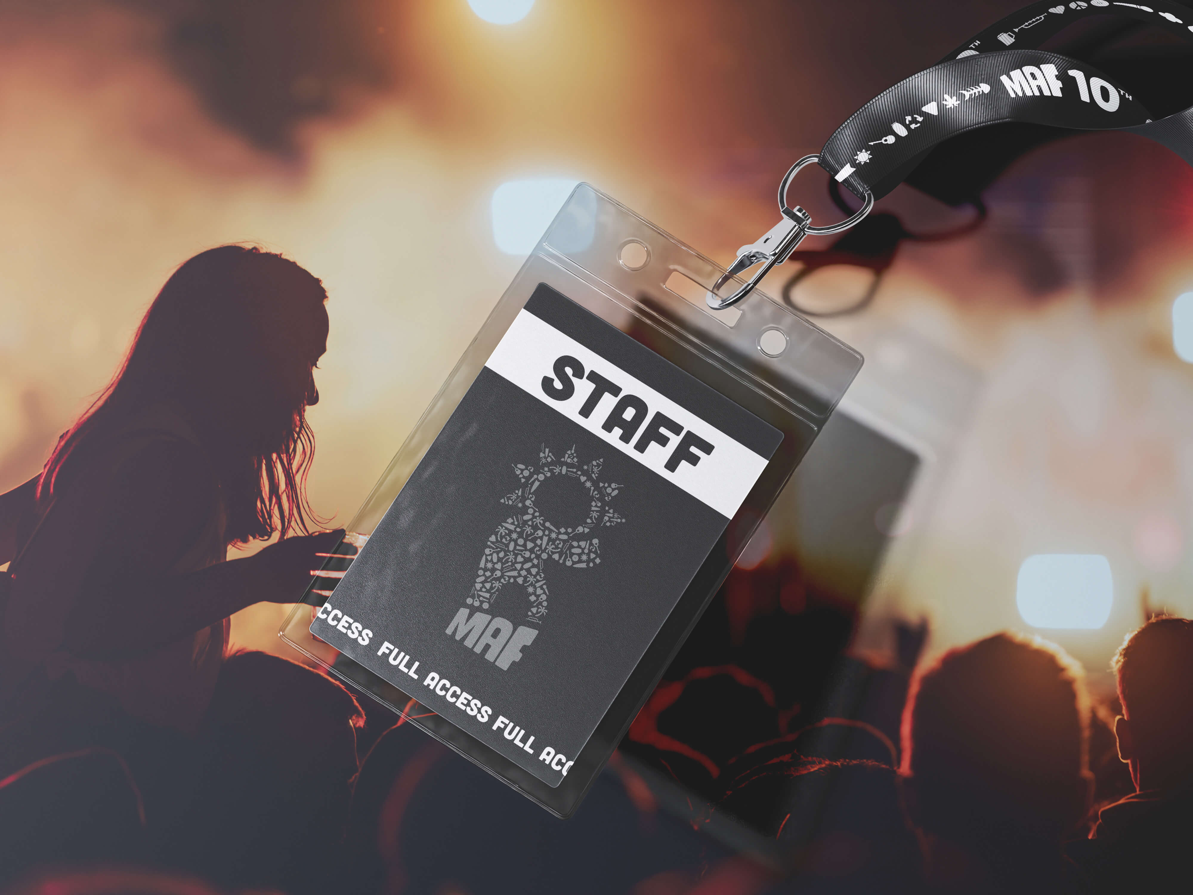 Black staff pass badge with MAF logo and white text reading 'STAFF' clipped to a black lanyard with white icons and 'MAF 10' text, over blurred background of a silhouetted crowd and stage with warm lighting.