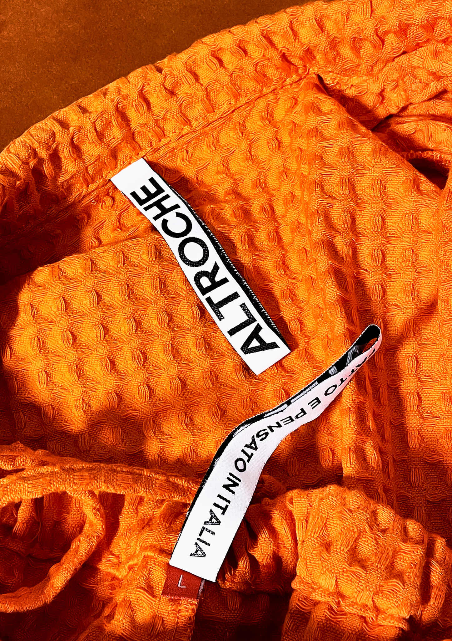 Close-up of textured orange fabric with two white labels: one says 'ALTROCHE' and the other partially visible says 'PENSATO IN ITALIA'.