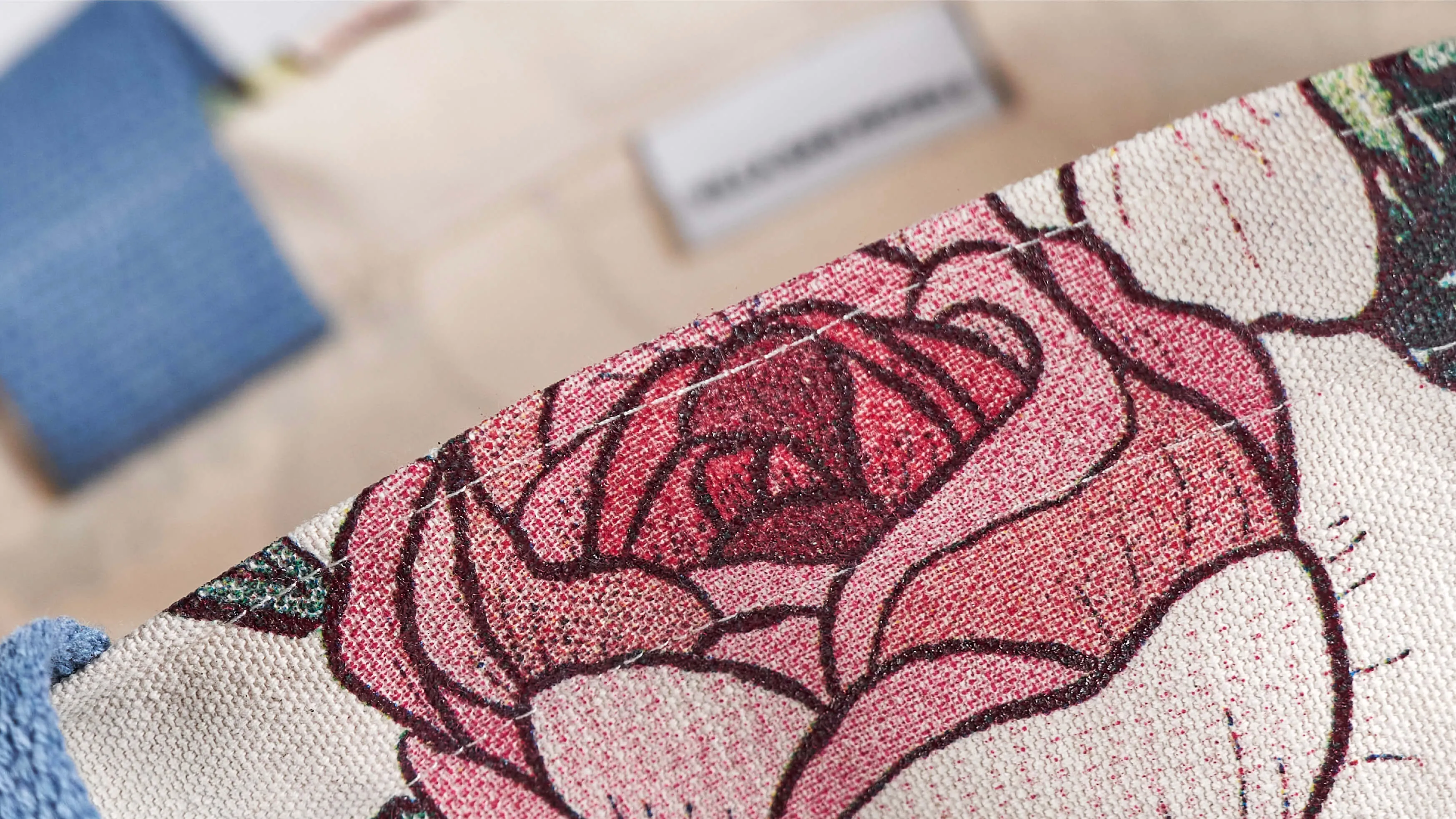 Close-up of tote bag corner with detailed pink and red rose pattern and textured fabric, showing part of a blue handle in the background.