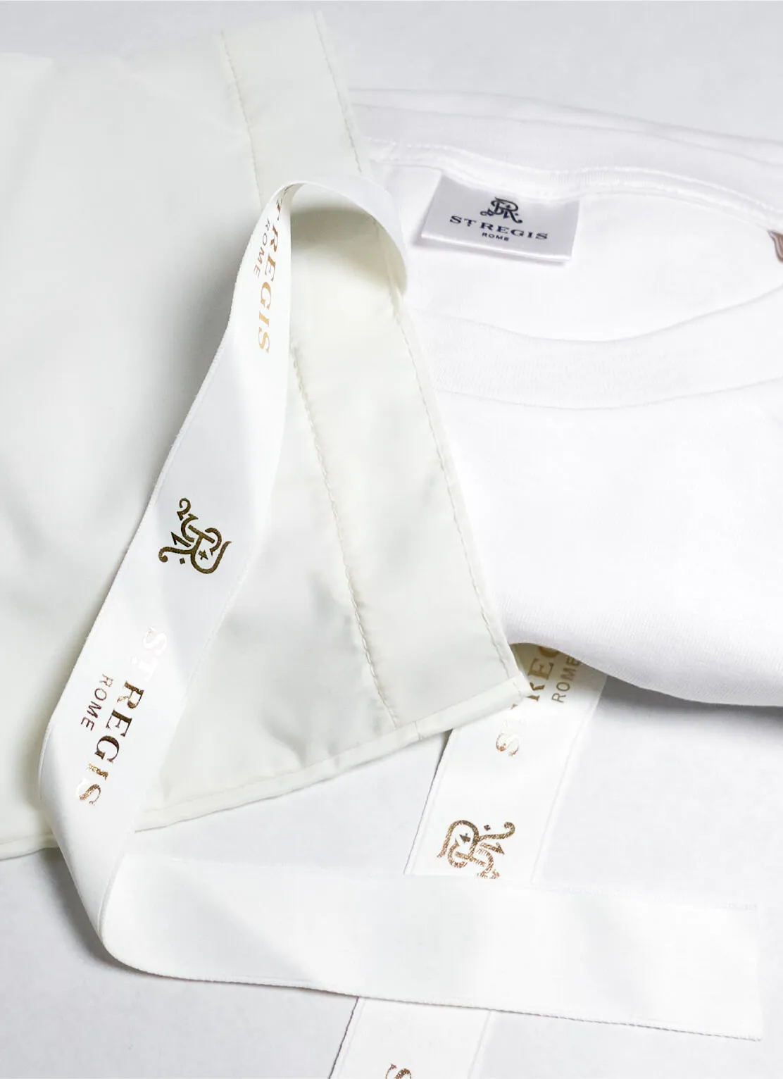 Close-up of a white folded t-shirt with a label inside the collar reading 'St Regis Rome' and a white satin ribbon with gold lettering and logo also reading 'St Regis Rome'.