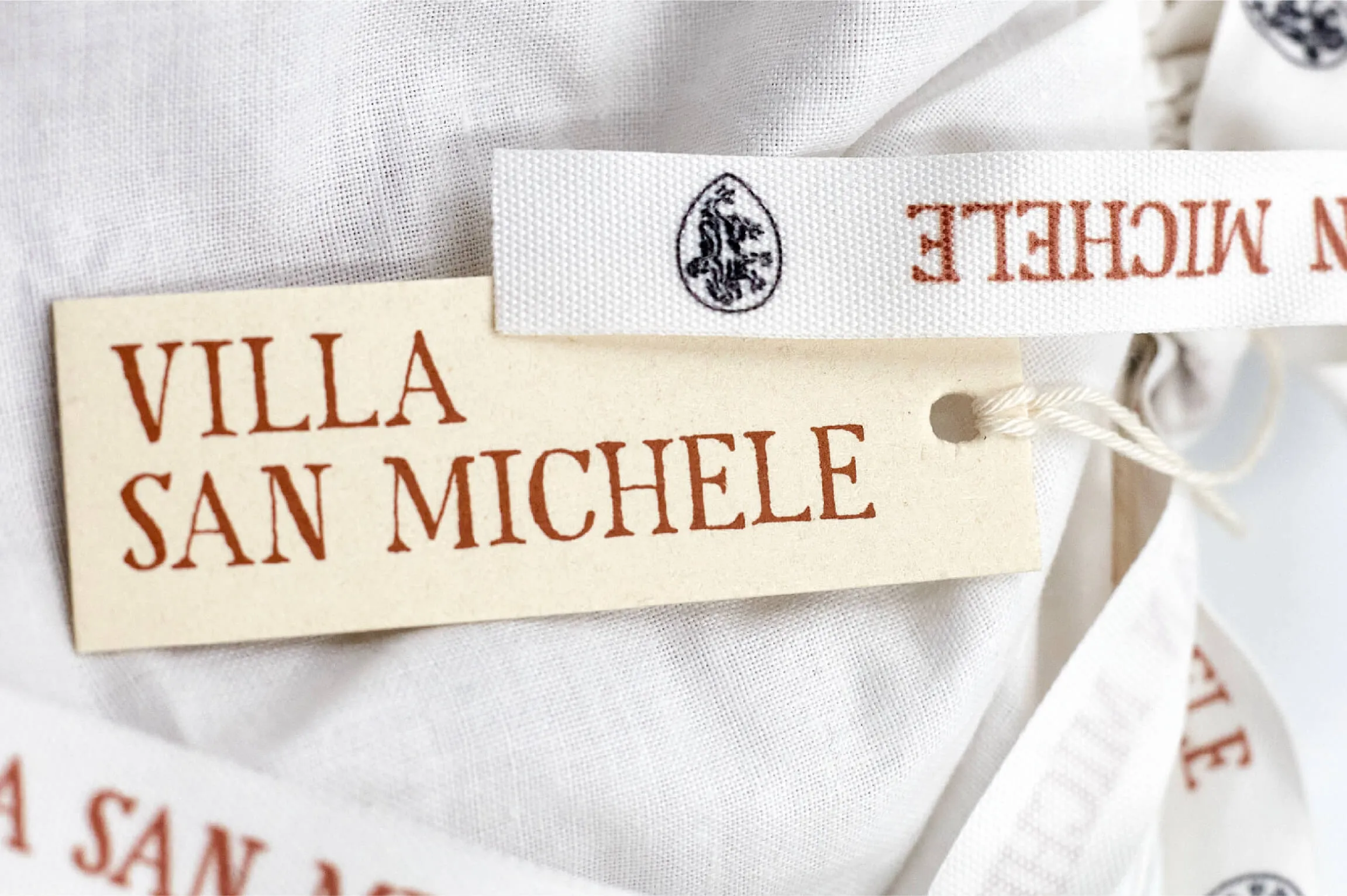 Close-up of a beige rectangular tag with 'VILLA SAN MICHELE' in bold reddish-brown letters attached with a white string to a textured white fabric with additional fabric labels featuring similar text and a small emblem.