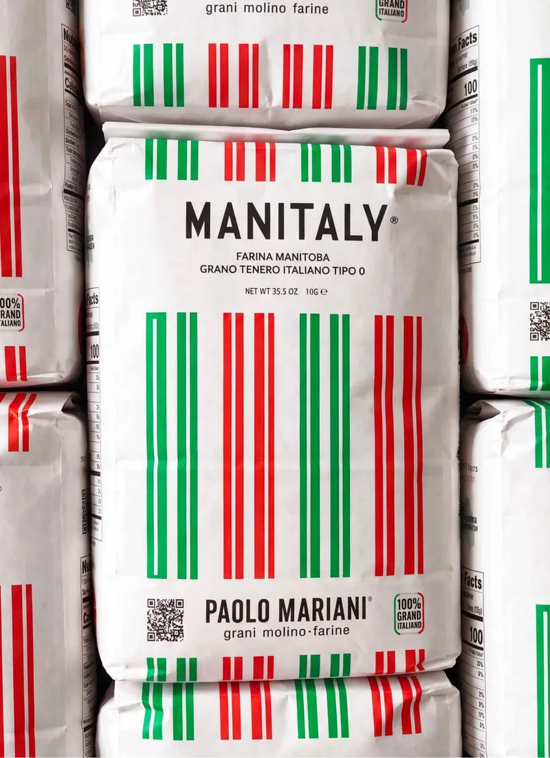 Stacked white bags of Manitaly Farina Manitoba Italian flour with green, white, and red barcode-like stripes, featuring Paolo Mariani branding and 100% Grano Italiano label.