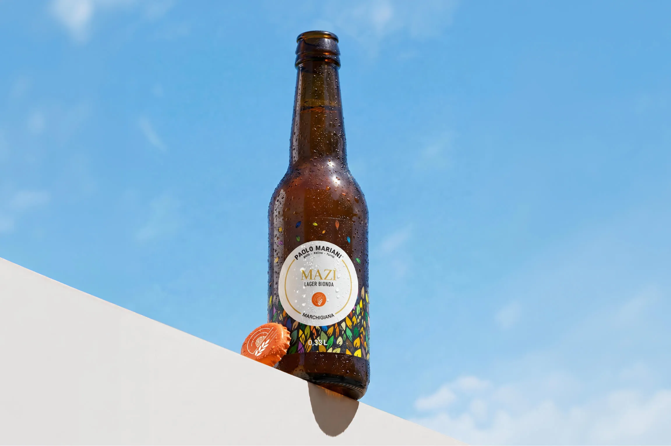 Brown beer bottle with condensation droplets standing on a white ledge against a blue sky, labeled 'Paolo Mariani Mazi Lager Bionda Marchigiana 0.33L', with an orange bottle cap resting beside it.