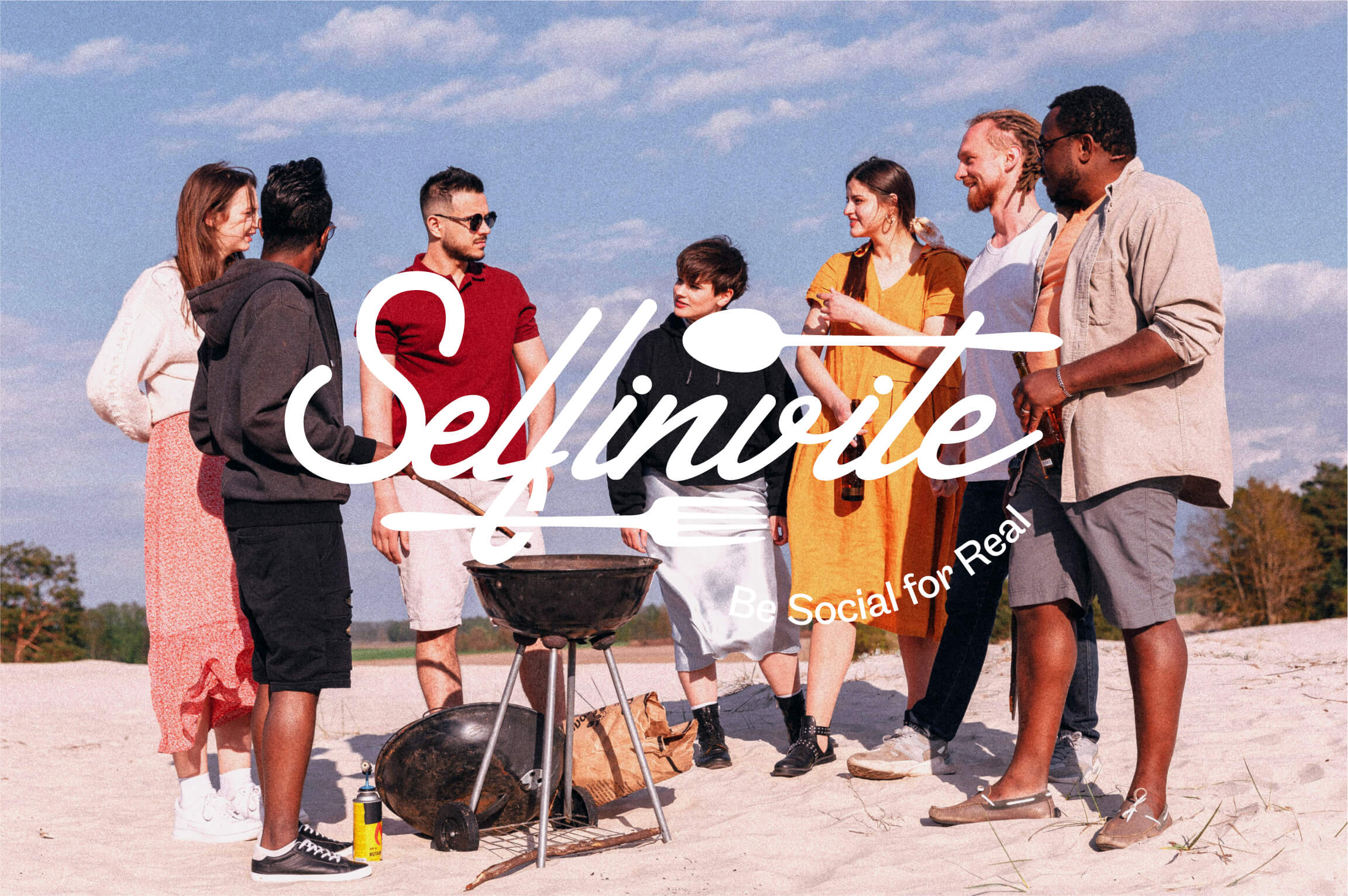 Seven diverse young adults standing on a beach with a charcoal grill and utensils, chatting and holding drinks under a partly cloudy sky.