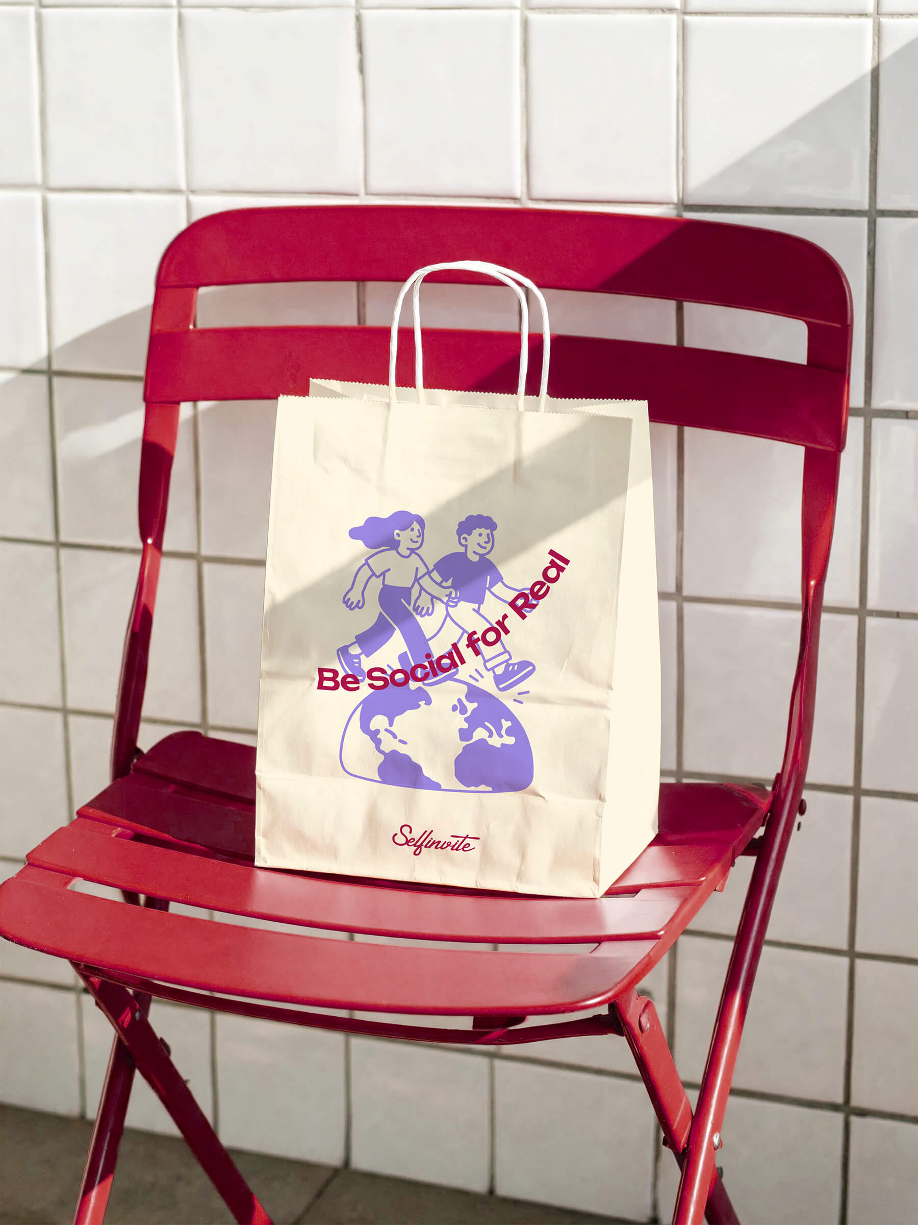 White paper shopping bag with illustration of two people walking on a globe, text 'Be Social for Real' and 'Selfinvite', placed on a red folding metal chair against a white tiled wall.