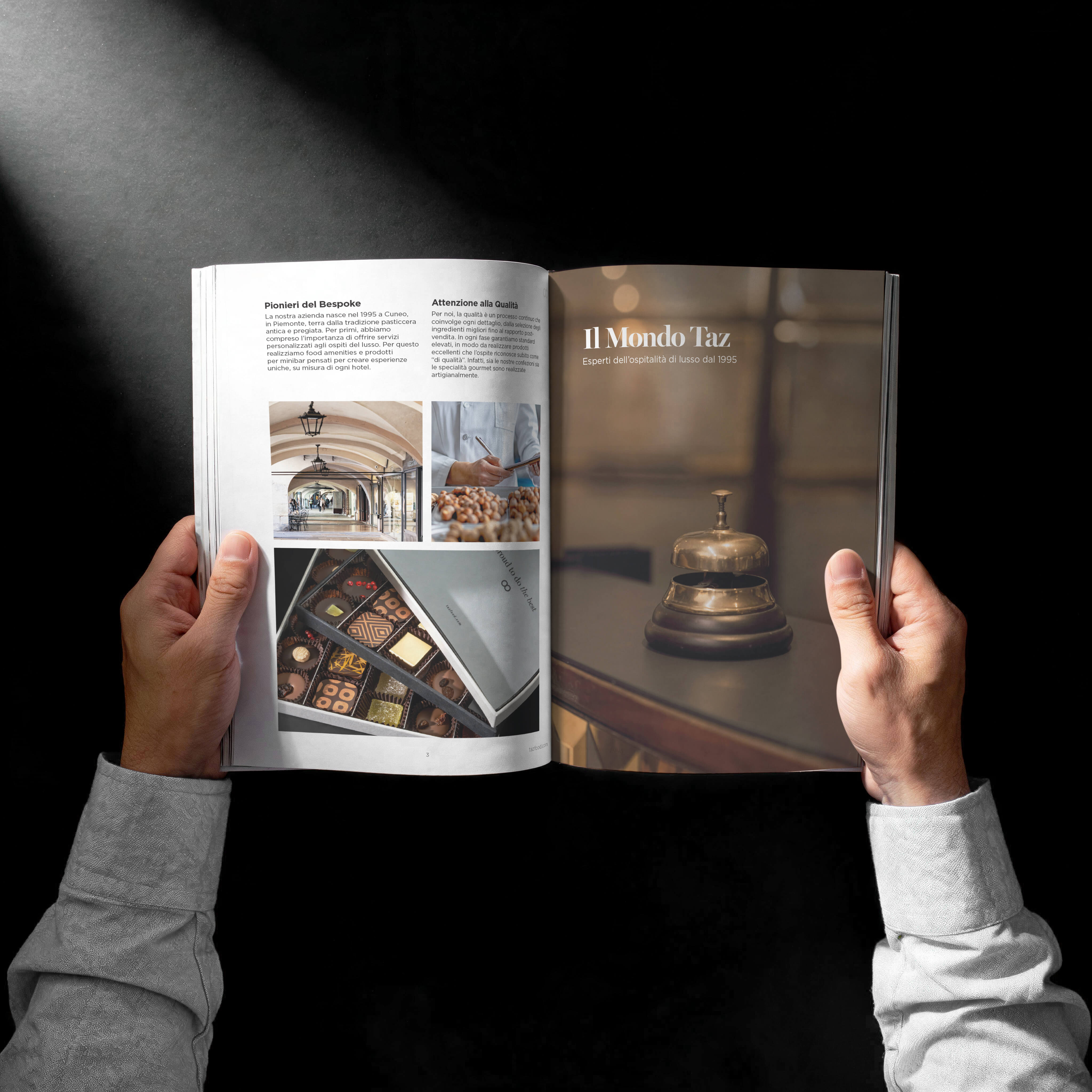 A person holding open a magazine or catalog featuring hospitality content; the left page shows Italian text with images of a covered walkway, a chef with pastries, and an open box of assorted chocolates, while the right page displays a hotel reception bell with the text "Il Mondo Taz Esperti dell'ospitalità di lusso dal 1995".