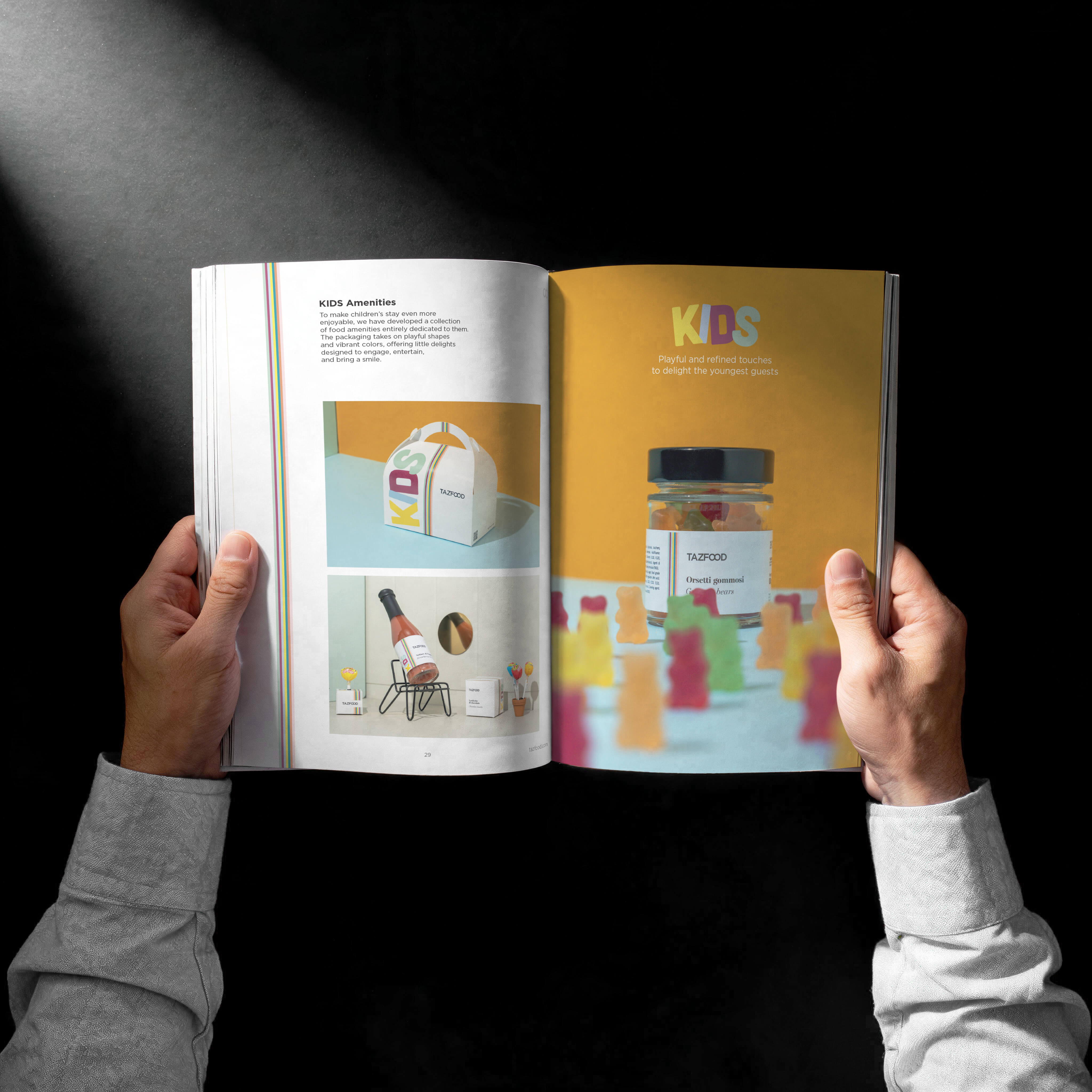 Hands holding an open magazine showing a two-page spread about KIDS amenities by TAZFOOD, featuring a colorful kids' food package and a jar of gummy bears with playful gummy bear shapes blurred in the foreground on a yellow background.